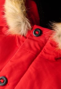 Czerwone okrycie wierzchnie z sztucznym futrem na kapturze, czarnym zapięciem na guziki i pikowaną strukturą. Materiał wydaje się gładki i wodoodporny.