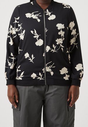 Person iført en sort jakke med lynlås og cremefarvet blomsterprint samt mørkegrå cargobukser, stående mod en ensartet lys baggrund.