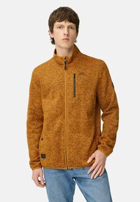 Orange fleecejakke med høj krave og lynlås, forsynet med sort lynlås lomme og logo detaljer, parret med blå jeans.
