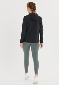 Kvinde med fletning, der går iført en sort hættetrøje, grønne leggings og hvide træningssko, på en helt hvid baggrund.