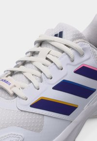 Κοντινό πλάνο λευκού αθλητικού παπουτσιού Adidas με λευκά κορδόνια και τρεις πολύχρωμες ρίγες σε ροζ, μπλε και κίτρινο στην πλαϊνή πλευρά.