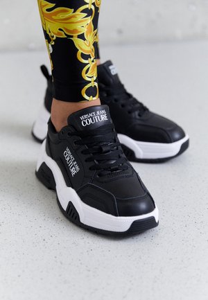 Sort-hvide Versace Jeans Couture sneakers, båret med sorte leggings med gule og gyldne barokmønstre.
