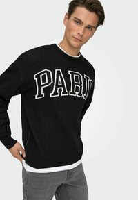 Pull en tricot noir avec l'inscription blanche audacieuse "PARIS" sur le devant, poignets et col côtelés, porté sur une chemise blanche.