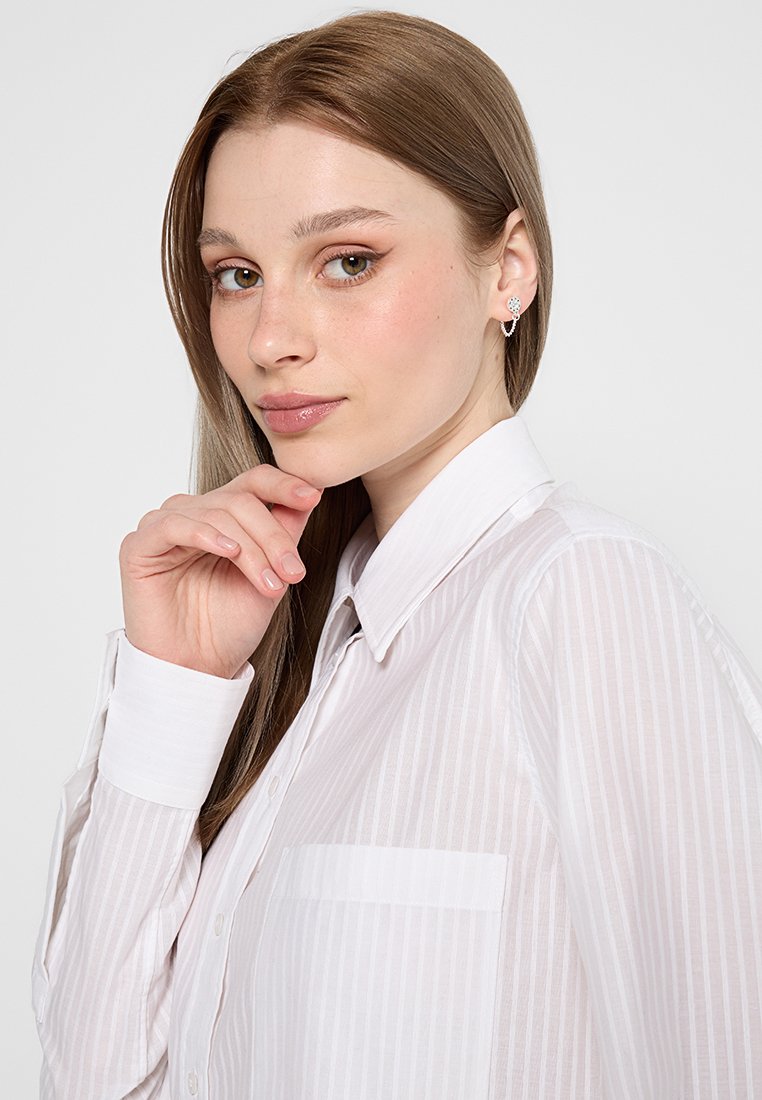 Biała, w paski koszula zapinana na guziki z kołnierzykiem i kieszenią na piersi, noszona przez modelkę z długimi brązowymi włosami. Gładka faktura, minimalistyczny design.
