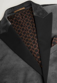 Grauer Samtblazer mit schwarzem Satin-Kragen. Futter zeigt ein geometrisches Muster in Braun auf Schwarz. Eine Taschenquadratik blitzt hervor.