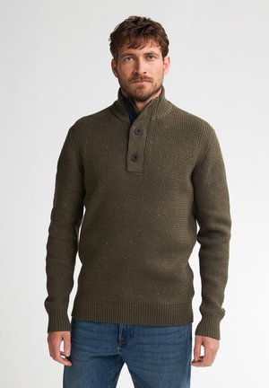 Man met bruin haar en baard, gekleed in een olijfgroen gebreid vest met knopen en een blauwe spijkerbroek, staand tegen een effen achtergrond.