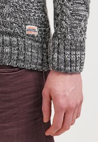 Sweter w splot sznurkowy w kolorze szarym z prążkowanym wykończeniem u dołu i na mankietach. Zawiera małą metkę z logo. Ręka spoczywa swobodnie na boku, ubrane w burgundowe spodnie.
