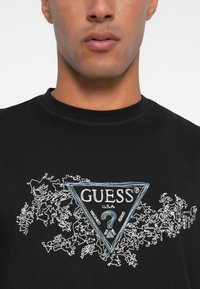 Must kõrvitsast valmistatud T-särk, millel on kolmnurkne sinine ja valge tikandiga logo tekstiga "GUESS®", täiendatud abstraktsete disainidega.