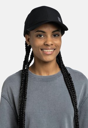 Junge Person mit langem, geflochtenem Haar, die eine schwarze Mütze und einen grauen Pullover trägt und vor einem einfarbigen hellen Hintergrund lächelt.