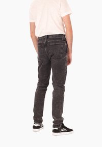 Homme portant un jean noir délavé, un t-shirt blanc et des baskets noir et blanc, se tenant de dos face à la caméra.