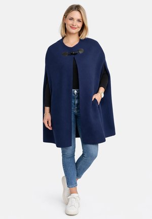 Donna che indossa un mantello di lana blu navy con fermaglio, maglia a maniche lunghe nera, jeans blu, sneakers bianche, in piedi con una mano in tasca.