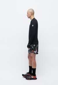 En person iført en svart langermet skjorte, blomstermønstrede shorts, svarte sokker og flerfargede joggesko, plassert på skrå.