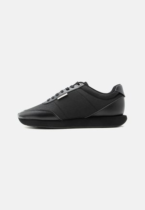 Schwarzer Sneaker in niedriger Ausführung mit strukturierten Seitenpaneelen, passenden schwarzen Schnürsenkeln und dicker, schräger Sohle auf weißem Hintergrund.