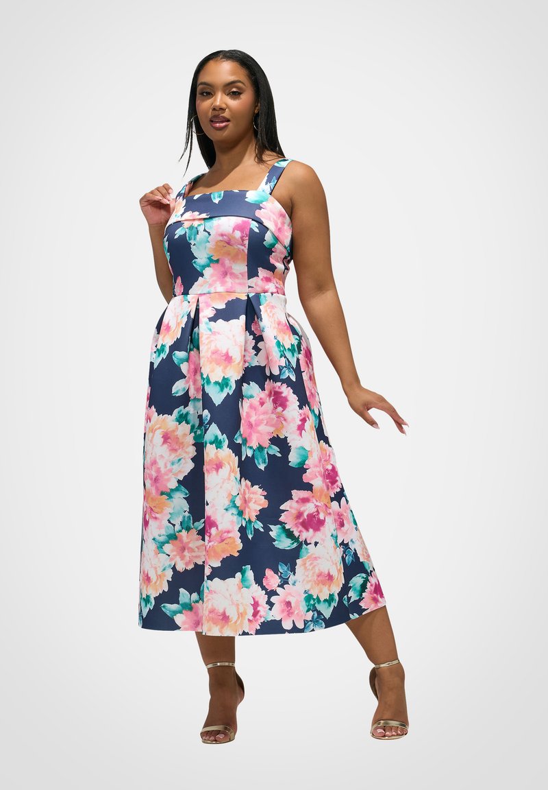 Frau trägt ein ärmelloses, marineblaues Midikleid mit großem rosa und türkisfarbenem Blumenmuster und posiert mit einer erhobenen Hand, während die andere nach unten ausgestreckt ist.