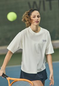 Jucătoare de tenis îmbrăcată într-un tricou alb din plasă, lejer, cu mâneci scurte, purtând o fustă închisă la culoare și ținând o rachetă portocalie.