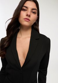 Blazer nero con scollatura a V profonda, spalle strutturate e tessuto testurizzato. Caratterizzato da un unico bottone di chiusura e vestibilità slim.