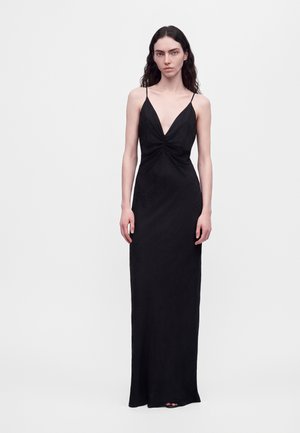 Un mannequin se tient devant un fond blanc uni, vêtu d’une robe noire longue jusqu’au sol, à fines bretelles et au décolleté en V plongeant.