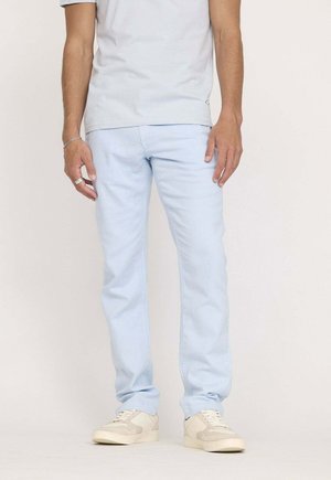 Pantalon en denim bleu clair, coupe slim, avec une texture lisse et des détails cousus. Associé à des baskets blanches.