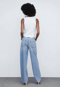 Haut blanc sans manches avec une texture côtelée, associé à un jean bleu clair à jambes larges. Le jean possède des poches arrière et une coupe décontractée.