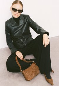 Veste en cuir noire, col roulé noir, large pantalon noir. La mannequin tient un sac à main marron à pois et porte des talons aiguilles marron assortis ainsi que des lunettes de soleil.