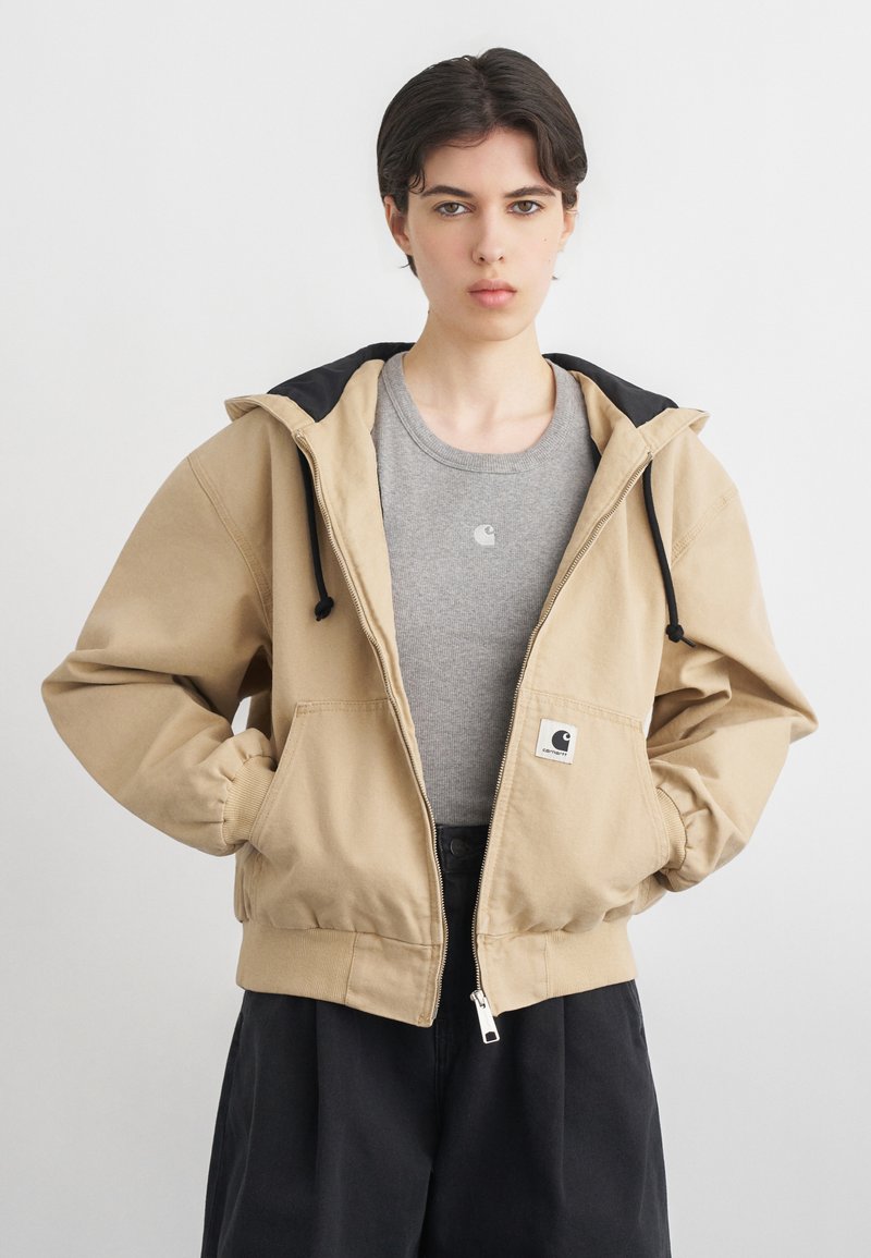 Personne aux cheveux courts et foncés portant une veste à capuche beige par-dessus un t-shirt gris avec logo et un pantalon noir, debout les mains dans les poches de la veste.