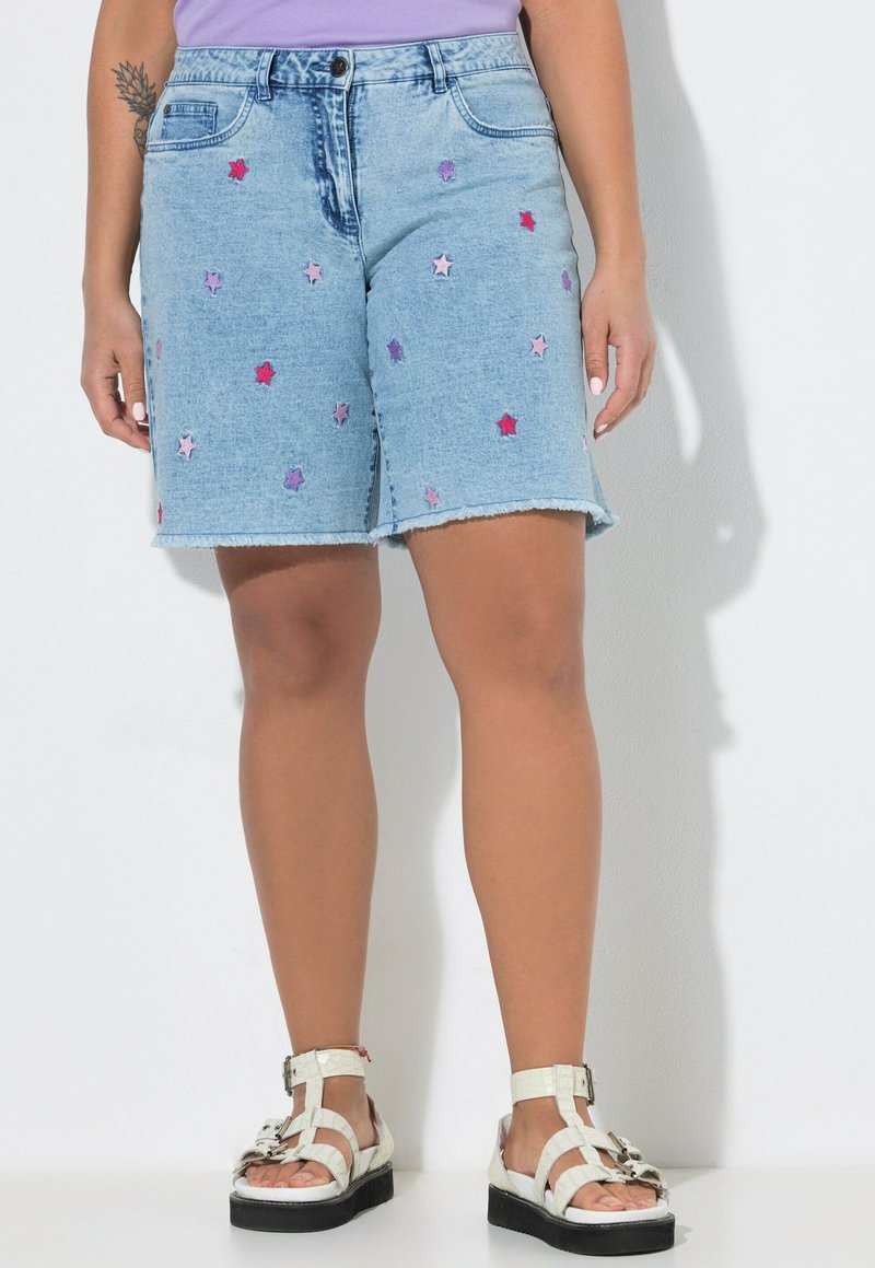 Shorts in denim celeste con stelle ricamate rosa e viola, indossati con sandali bianchi e una maglia lilla, visibili dalla vita ai piedi.