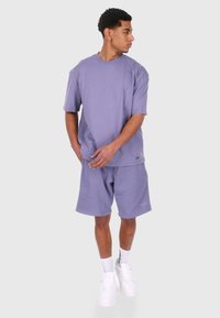 Lys lilla oversized t-skjorte og matchende shorts i et mykt, bomullsliknende materiale; har subtil merkevare på shortsene, kombinert med hvite sneakers.