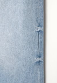 Tissu en denim bleu clair montrant une couture verticale avec un léger délavage et des plis le long du bord droit sur un fond blanc.