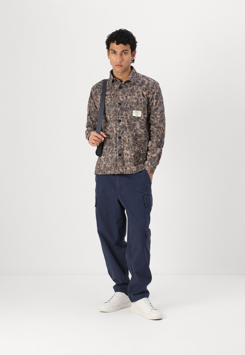 Chemise à manches longues avec un motif paisley marron et bleu marine, associée à un pantalon cargo bleu marine et des baskets blanches. Sangle noire incluse.