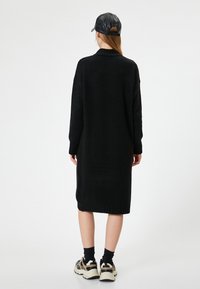 Abito maglione nero fino al ginocchio realizzato in tessuto a maglia. Presenta un colletto alto e polsini a coste, abbinato a sneaker chunky.