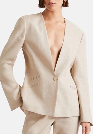Blazer beige su misura con profondo scollo a V e chiusura con un solo bottone, indossato con pantaloni a vita alta coordinati.