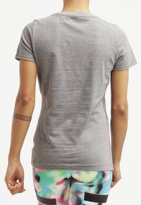 Osoba zobrazená zezadu, oblečená v šedé košili s krátkým rukávem s malými perforacemi a barevných legínách s abstraktním vzorem.