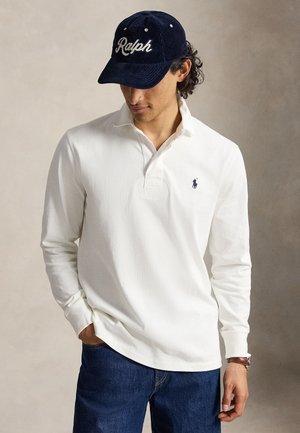 Homme portant un polo blanc à manches longues avec un petit logo, un jean bleu, une casquette bleu marine avec l'inscription Ralph, et une montre-bracelet, regardant vers le bas.