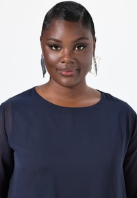 Marineblaue Bluse mit kurzen, transparenten Ärmeln. Auffällige silberne Ohrringe in geschwungenem Design. Glatte Stoffstruktur und runder Ausschnitt.