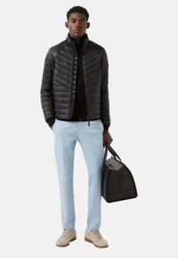 Černá prošívaná bunda s dlouhými rukávy a zapínáním na zip, v kombinaci s černou košilí, světle modrými kalhotami a béžovou obuví. Drží černou tašku.