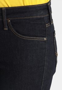 Jeans de mezclilla oscura con un diseño ajustado, que presentan costuras naranjas, dos bolsillos y un ligero estiramiento para mayor comodidad.