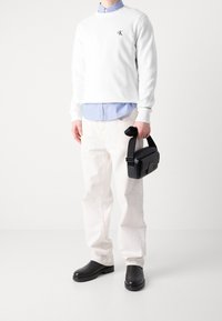 Vit bomullströja med CK-logotyp, blårandig skjorta under, off-white byxor, svart läderväska med rem och svarta skor.