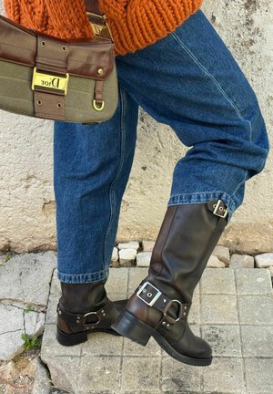 Persona che indossa jeans blu, stivali in pelle marrone scuro con fibbie, maglione arancione a maglia e porta una borsa Dior marrone e verde oliva.