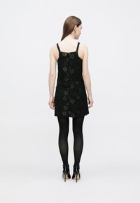 Juoda suknelė su tekstūruota paviršiaus danga, puošta gėlių aplikacijomis, plonomis petnešėlėmis ir užsegimu už nugaros. Dėvima su juodomis kojinėmis ir aukštakulniais.