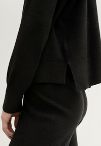 Maglione nero in morbido filato, con una fessura laterale e una vestibilità rilassata. La texture è soffice, con maniche lunghe e orlo arrotondato.