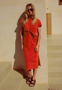 Rotes, mittellanges Kleid mit einem geknoteten Design an der Vorderseite. Kurze Ärmel, glatte Textur. Getragen mit schwarzen Sandalen mit goldenen Schnallen.