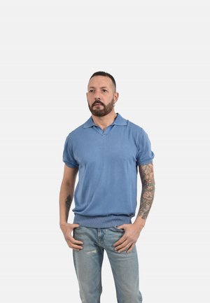 Uomo con barba e tatuaggi in piedi con le mani nelle tasche dei jeans, indossa una polo blu a maniche corte su sfondo semplice.