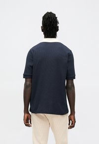 Laivastonsininen lyhythihainen poolopaita, jossa on teksturoitu neulostyyli ja kermaisen värinen kaulus; yhdistetty vaaleisiin kuviollisiin housuihin.