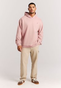 Lichtroze hoodie met een voorzak, voorzien van een klein logo op de borst. Draagt met beige broek en bruine sneakers. Gladde textuur.