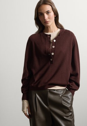 Mujer con cabello largo y castaño viste un suéter marrón oscuro con botones sobre una camiseta beige y pantalones de cuero oscuro, de pie contra un fondo claro.