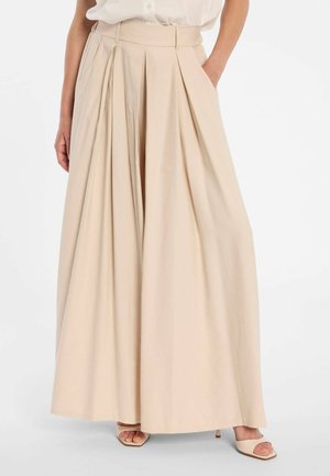 Frau trägt hochtaillierte, weit geschnittene beigefarbene Hose mit Falten, kombiniert mit beigen offenen Sandaletten mit Absatz und einem in die Hose gesteckten weißen Hemd.