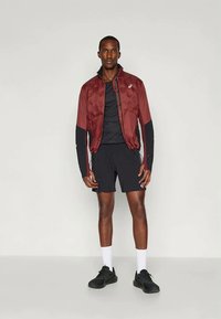 Giubbotto leggero bordeaux con fronte in tessuto strutturato, maniche nere, maglietta sportiva nera, pantaloni corti neri e sneakers nere. Calzini bianchi in vista.