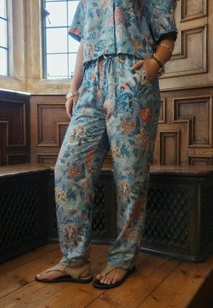 Person wearing loose, floral-patterned blue pants and matching short-sleeve shirt, standing barefoot on wooden floor by window and wooden paneling.