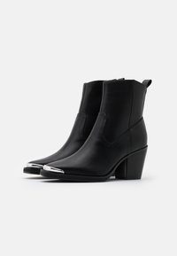 Botas de cuero negro hasta el tobillo con punta afilada, detalles metálicos plateados en la punta y un tacón apilado. Cuenta con lengüetas en la parte trasera.