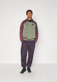 Zielona i fioletowa kurtka z półzamek z czerwonymi paskami, zestawiona z fioletowymi spodniami. Posiada elastyczne mankiety, kieszenie i akcenty z logo. Czarne sneakersy.